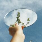 Hand holding a mirror reflecting sky and leaves, showcasing creativity and nature.